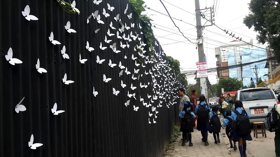 a wall by a pavement filled with paper-cut white butterflies with people passing by looking at the wall