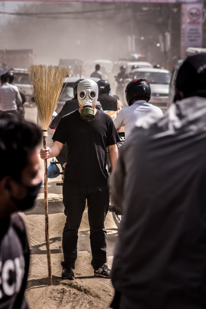 a civilian man standing in the street wearing a gas mask and holing a brush in his right arrm, while motorcyclists pass by